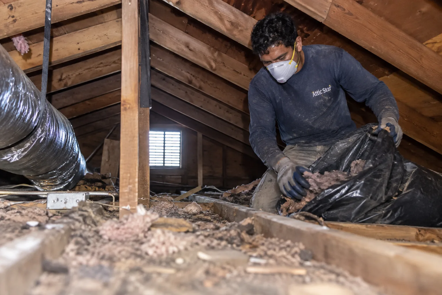 A male technician wearing a dust mask and gloves inspects and removes debris from a residential attic in an older San Francisco home, showing realistic signs of dust and wear on his clothing.