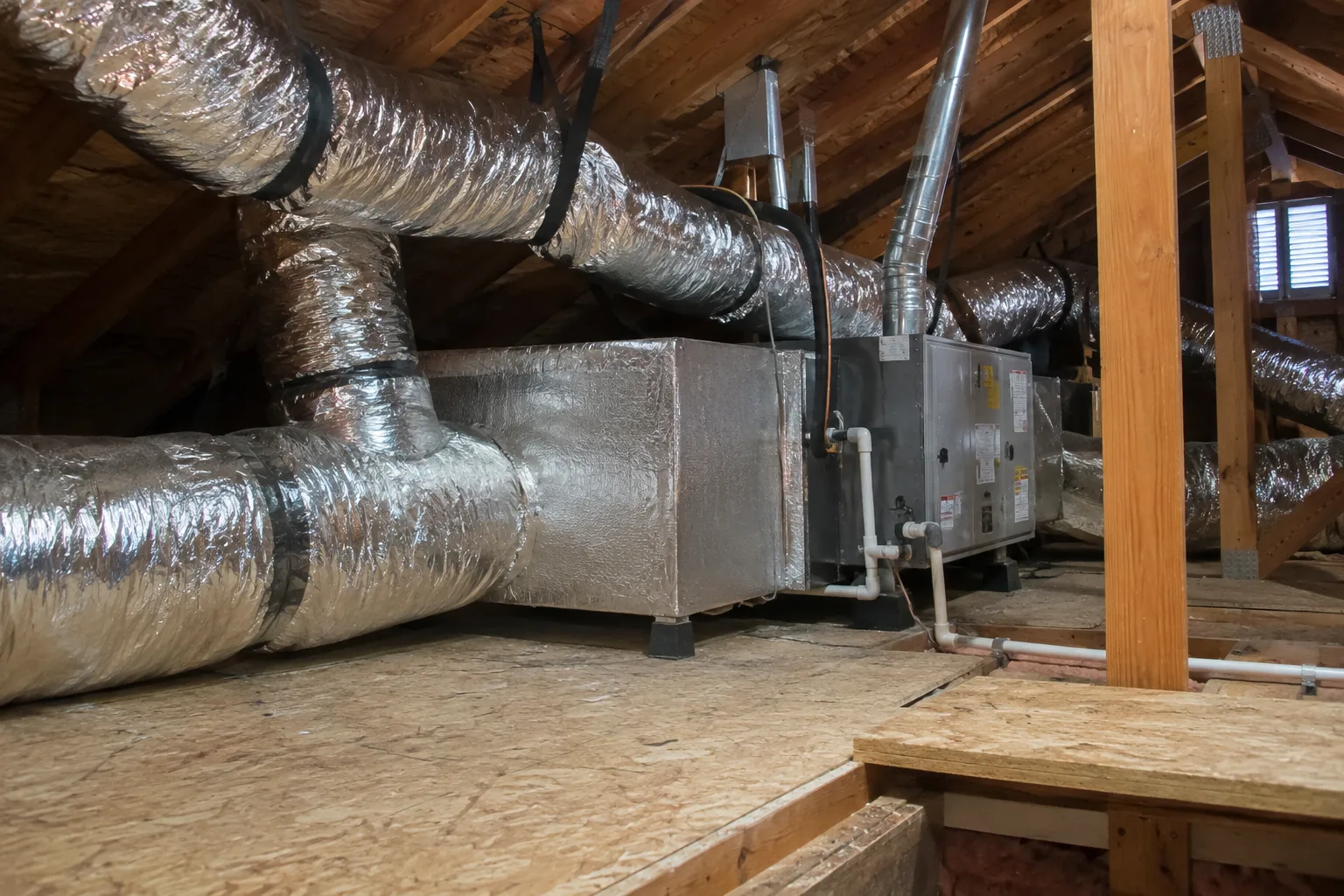 Attic furnace connected to insulated supply and return ducts, set on a plywood platform beside the attic walkway.